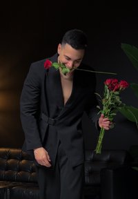 Fekete elegáns öltöny ezüst lánccal, vörös rózsákat tartva. Az öltöny szabott kialakítással és mély V-alakú nyakkivágással rendelkezik. Sötét háttér.