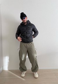 Homme portant un bonnet noir, des lunettes de soleil foncées, une doudoune noire, un pantalon ample olive et des baskets blanches, debout à l'intérieur contre un mur blanc.