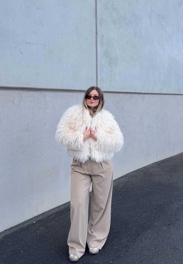 Femme portant une veste blanche duveteuse surdimensionnée, un pantalon beige à jambes larges, des lunettes de soleil, debout sur l'asphalte près d'un mur en béton gris.