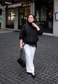 Fekete dzsekit és fehér nadrágot viselő nő, aki fekete táskával és napszemüveggel sétál egy köves utcán, egy bolt előtt.