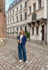 Veste portefeuille bleu marine portée sur un haut blanc, associée à un jean large foncé et des baskets blanches. Rue pavée et bâtiments historiques en arrière-plan.