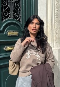 Pullover aus hellbraunem Wolle mit Hundemotiv; grauer Faltenrock; hält eine Brille; beiger Tasche; steht neben einer grünen Tür mit verzierten Details.