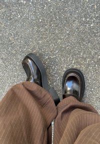 Błyszczące czarne buty na platformie noszone z brązowymi spodniami w prążki, stojące na chropowatej powierzchni asfaltu.