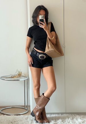 Schwarzes, eng anliegendes T-Shirt, hochgeschnittene schwarze Shorts, braune Lederstiefel bis zum Knie, silberne Gürtelschnalle, beigefarbene Tragetasche, verspiegelt im Hintergrund.
