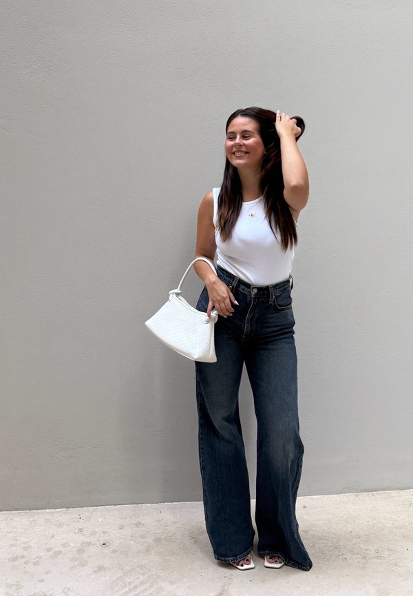 Mujer con camiseta sin mangas blanca y vaqueros oscuros de pierna ancha, sosteniendo un bolso blanco, sonriendo y tocándose el pelo frente a una pared gris lisa.