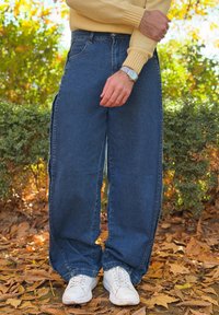 Άτομο που φοράει φαρδιά μπλε τζιν, λευκά αθλητικά παπούτσια, κίτρινο πουλόβερ και ρολόι στον καρπό, στέκεται πάνω σε πεσμένα φθινοπωρινά φύλλα σε εξωτερικό χώρο.