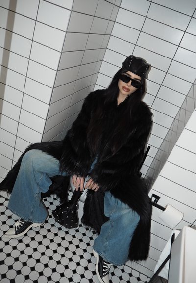 Persona con un abrigo de piel sintética negra, vaqueros de pernera ancha, sombrero negro y gafas de sol, sentada en un inodoro en un baño con azulejos.