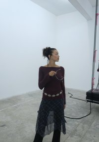 Young woman with curly hair styled in a bun, dressed in a burgundy top and a plaid skirt, holding sunglasses in a minimalist room featuring swing seats.