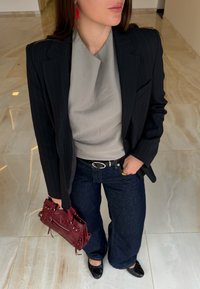 Czarna marynarka w prążki na jasnoszarym topie, ciemnoniebieskie dżinsy w stylu dzwonów, czarne baleriny, trzymając torebkę w kolorze buraczkowym o fakturze. Widoczne czerwone kolczyki.