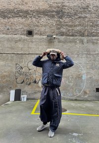 Námořnická parka s kapucí podšitou kožešinou, předními kapsami, v kombinaci s volnými černými kalhotami a šedými kostkovanými slip-on botami v městském prostředí.