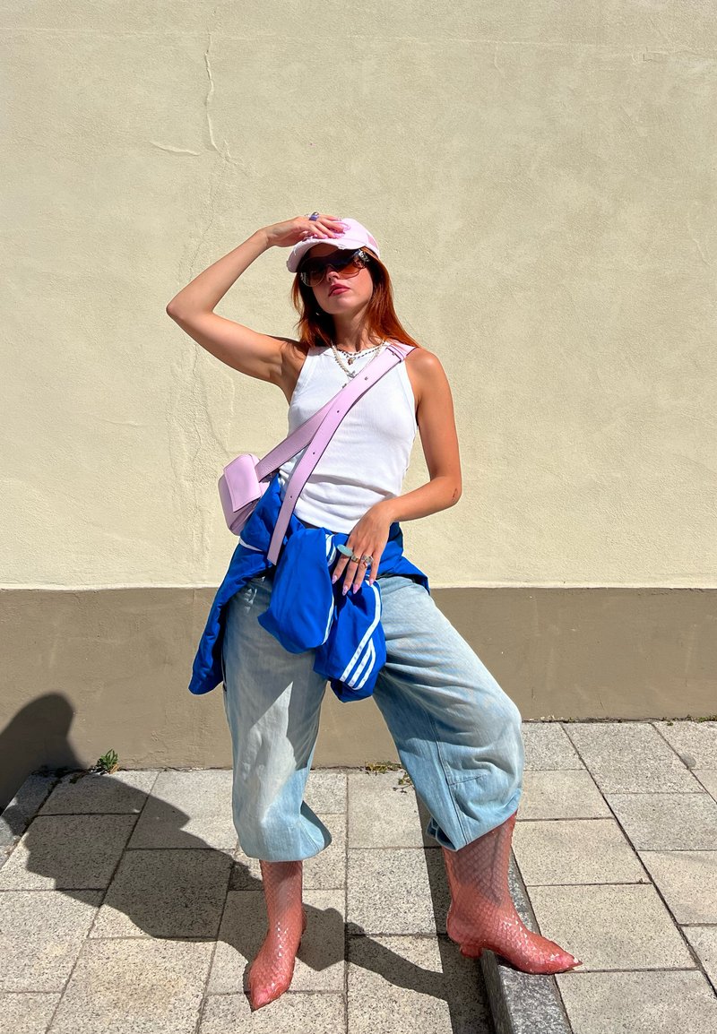 Μοντέλο φορώντας λευκή αμάνικη μπλούζα, ανοιχτού γαλάζιου τζιν και ροζ δίχτυνα μποτάκια, με ροζ καπέλο και τσάντα ώμου, στέκεται σε εξωτερικό χώρο μπροστά από έναν ουδέτερο τοίχο.