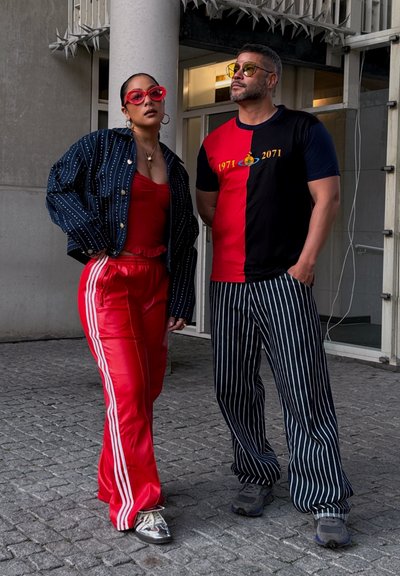 Dos personas están de pie en una acera. Una lleva una chaqueta de rayas azul marino, una camiseta sin mangas roja y pantalones de chándal rojos con rayas blancas. La otra tiene una camiseta dividida en negro y rojo y pantalones a rayas. Ambas llevan gafas de sol.