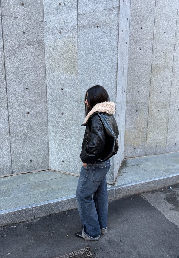 Schwarze Lederjacke mit Lammfellkragen, kombiniert mit lockeren blauen Jeans und spitzen schwarzen Schuhen, stehend vor einer strukturierten Steinmauer.