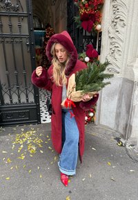 Manteau à carreaux rouge et noir avec une capuche bordée de fourrure, jeans larges bleus et chaussures rouges, tenant une branche de sapin enroulée. Feuilles jaunes au sol.