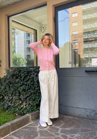 Ανοιχτό ροζ κασκόλ με πλέξη καλωδίου και κουμπιά, συνδυασμένο με φαρδιές λευκές παντελόνες. Στέκεται κοντά σε μια γυάλινη πόρτα με πράσινο φόντο.