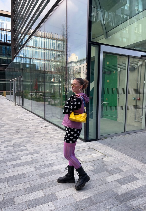 Naine, kes kannab täpilist topi, lilla vestit, triibulisi retuuse, musti saapaid ja kollast kotti, seisab käed rüpes kaasaegse klaashoone ees.