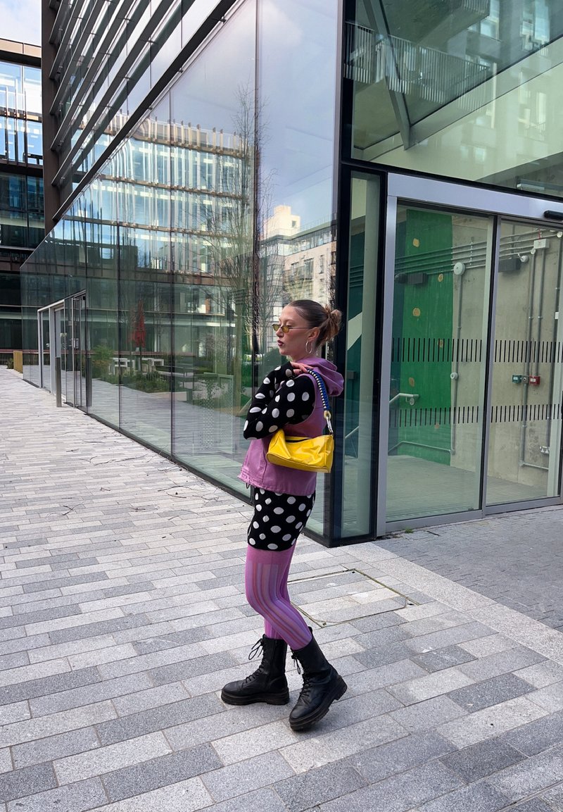 Žena u točku topu, ljubičastom prsluku, prugastim leginsama, crnim čizmama i žutom torbom stoji s preklopljenim rukama ispred moderne staklene zgrade.