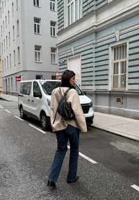 Beige bunda, černá volná taška a rozšířené džíny na osobě, která jde po ulici ve městě poblíž b�ílé dodávky a budovy s modrými pruhy.