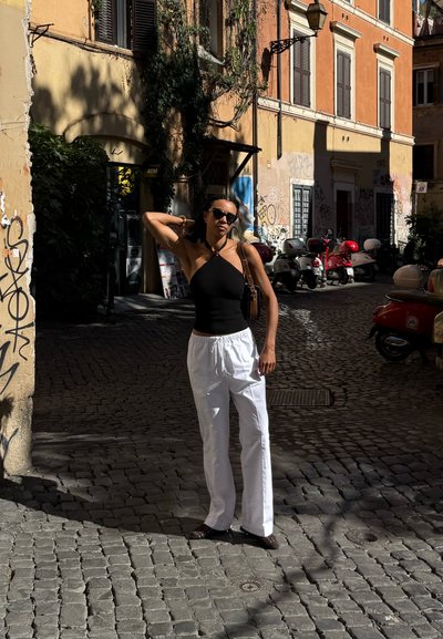 Top negro de halter, pantalones blancos de tiro alto y gafas de sol. De pie en una calle empedrada cerca de paredes cubiertas de grafitis y scooters aparcados.