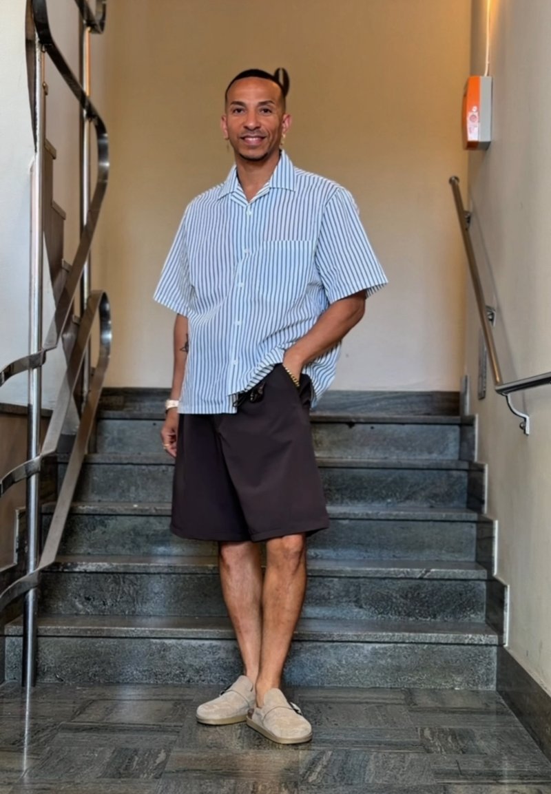 Homme debout dans un escalier intérieur, portant une chemise à manches courtes rayée, un short foncé et des chaussures légères sans lacets, souriant avec une main dans la poche.