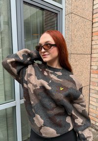 Camisola com padrão camuflado em mistura de lã cinzenta, castanha e bege, com decote redondo e ombros caídos, combinada com óculos de sol castanhos de armação retangular.