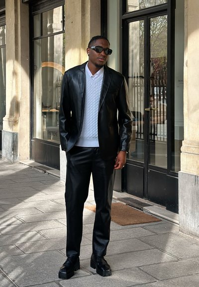 Hombre con blazer y pantalones de cuero negro, suéter blanco de punto tipo trenza y gafas de sol negras, de pie sobre un pavimento de piedra frente a puertas de cristal.