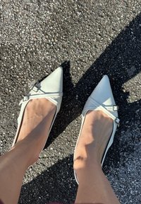 Vita spetsiga ballerinaskor i slät material, med en enkel remdetalj som har ett silverfäste. Platt sula, minimalistisk design.