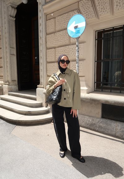 Chaqueta de gran tamaño en verde oliva con botones negros, pantalones anchos negros y un bolso de mano negro tejido. Gafas de sol grandes y negras y un hijab.
