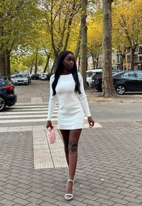 Valkoinen pitkähiha mini-mekko, yhdistetty vaaleanpunaiseen clutchiin. Jalassa vaaleansiniset korolliset sandaalit. Taustalla syksyisiä puita ja pysäköityjä autoja.