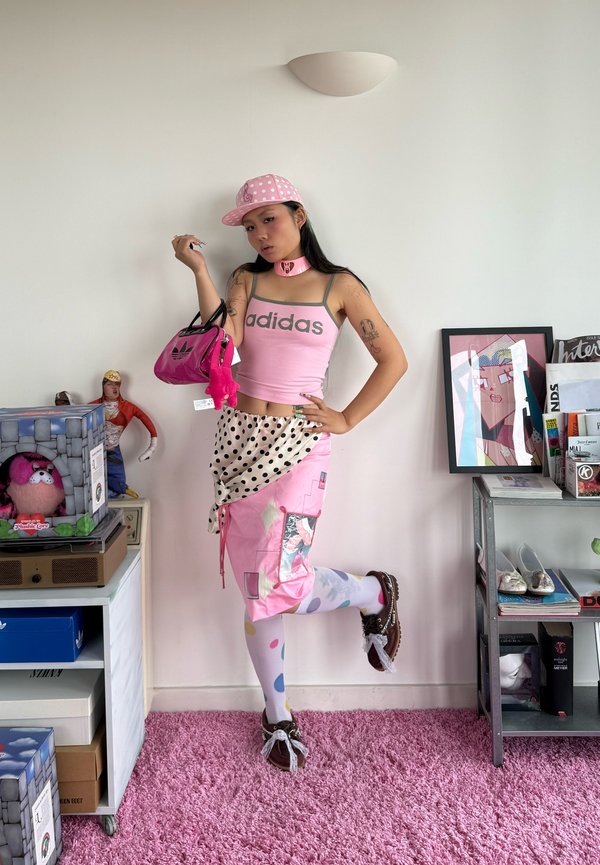 Nuori nainen, joka pukeutunut vaaleanpunaiseen Adidas-hihattomaan paitaan, pallokuvioiseen hameeseen ja värikkäisiin sukkiksiin, poseeraa pitäen vaaleanpunaista laukkua huoneessa, jossa on leluja ja kehystettyä taidetta.