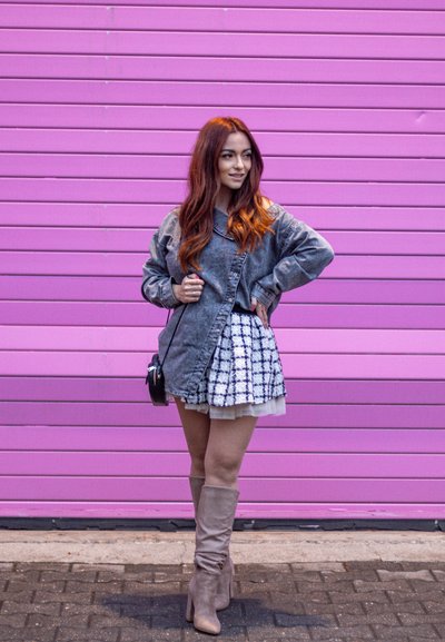 Chaqueta de denim oversized gris, falda blanca texturizada con patrón de cuadros negros, botas beige hasta la rodilla y un pequeño bolso cruzado negro. Fondo rosa.