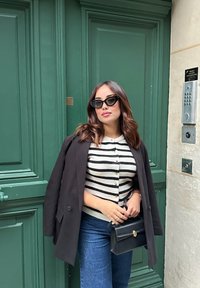 Schwarze Blazer über einem creme- und schwarz gestreiften Cardigan, blaue gerade Jeans, hält eine schwarze Handtasche; Hintergrund mit grüner Tür.