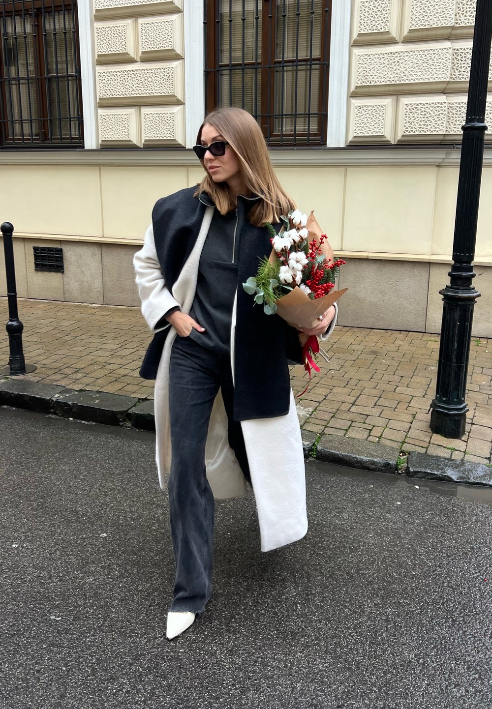 Femme en manteau noir et blanc et lunettes de soleil, marchant dans la rue, tenant un bouquet de coton et de baies rouges enveloppé dans du papier kraft brun.