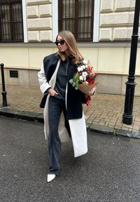 Femme en manteau noir et blanc et lunettes de soleil, marchant dans la rue, tenant un bouquet de coton et de baies rouges enveloppé dans du papier kraft brun.