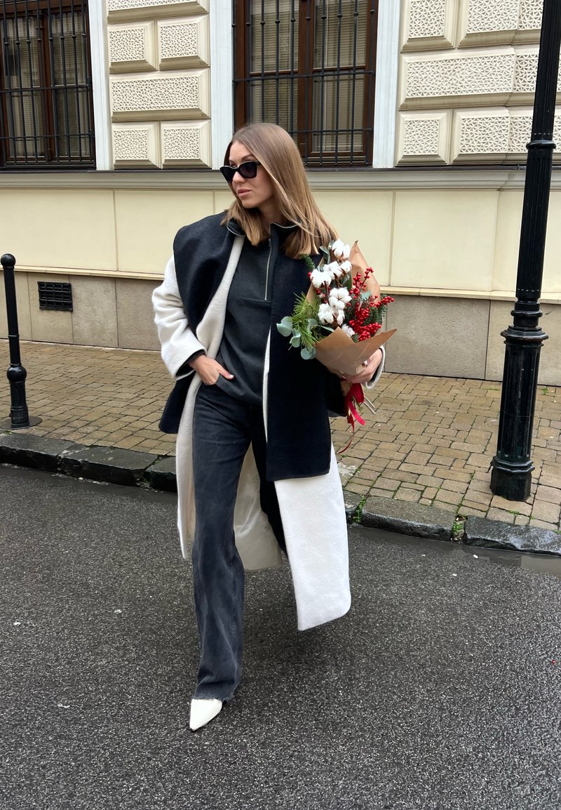 Ženska v črno-beli jakni in sončnih očalih hodi po ulici, v roki drži šopek bombaža in rdečih jagod, zavitih v rjavi papir.