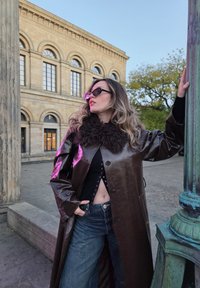 Lesklý hnedý kožený kabát s huňatým golierom, štýlovo oblečený cez čierne tričko a voľné modré denimové džínsy, doplnený okrúhlymi slnečnými okuliarmi.