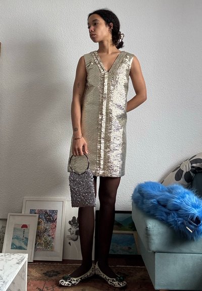 Persona con un vestido sin mangas dorado brillante, medias negras, sosteniendo un bolso de lentejuelas plateado, de pie junto a un sofá azul con un abrigo de piel azul.