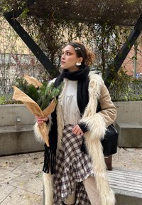 Cappotto in finta pelliccia tan con accenti bianchi, sciarpa nera, blusa bianca e gonna a quadri. Tiene un bouquet di verde avvolto in carta marrone.