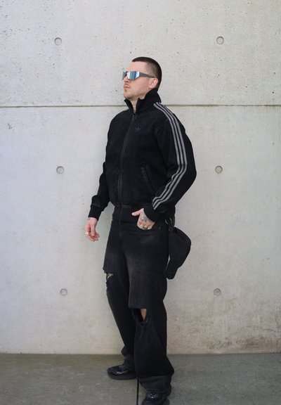 Chaqueta de mezclilla negra con rayas grises, llevada sobre jeans negros desgastados. Los accesorios incluyen gafas de sol reflectantes y un pequeño bolso negro.