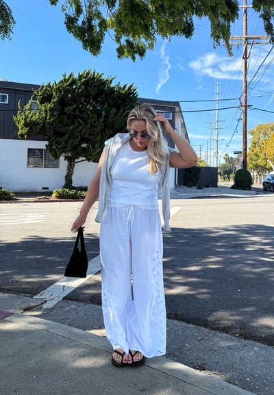 Mujer con top blanco sin mangas y pantalones anchos se arregla el cabello, sosteniendo un bolso negro, de pie en una esquina soleada de la calle bajo la sombra de un árbol.
