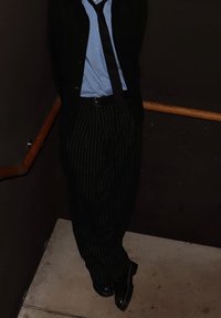 Pantaloni cu dungi negre, cămașă bleu deschis, cardigan negru și cravată, stând pe o scară pe fundal întunecat. Pantofi negri vizibili.