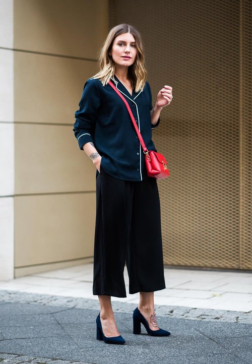 Fekete, bő szárú nadrág, sötétkék, gombos ing fehér szegélyezéssel, piros keresztpántos táska, kék magassarkú cipő, áll a köves utcán.