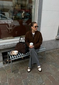 Καφέ σουέντ μπουφάν, γκρι φαρδιές παντελόνες, λευκοκάστανα loafers, στρογγυλά γυαλιά ηλίου, κράτησε ένα φλιτζάνι καφέ. Μαύρη τσάντα πάνω σε μια παγκάκι.