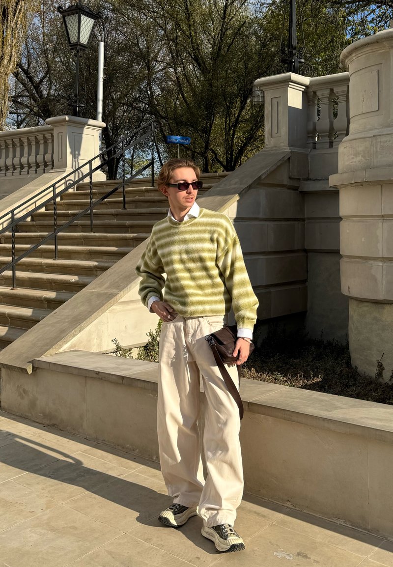 Ein junger Mann mit Sonnenbrille, grün gestreiftem Pullover, weißen Hosen und Sneakers steht in einer sonnigen Außenumgebung in der Nähe von Steintreppen und einem Geländer.