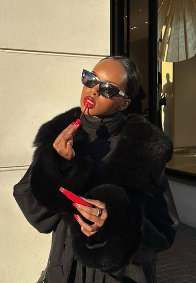 Mujer con abrigo de piel negro y gafas de sol se aplica un brillante lápiz labial rojo al aire libre bajo la luz del sol cerca de la entrada de un edificio.