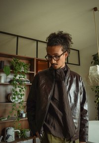 Mann med krøllete hår og briller, iført en mørkebrun skinnjakke og skjerf, som står innendørs nær en trehylle med planter.