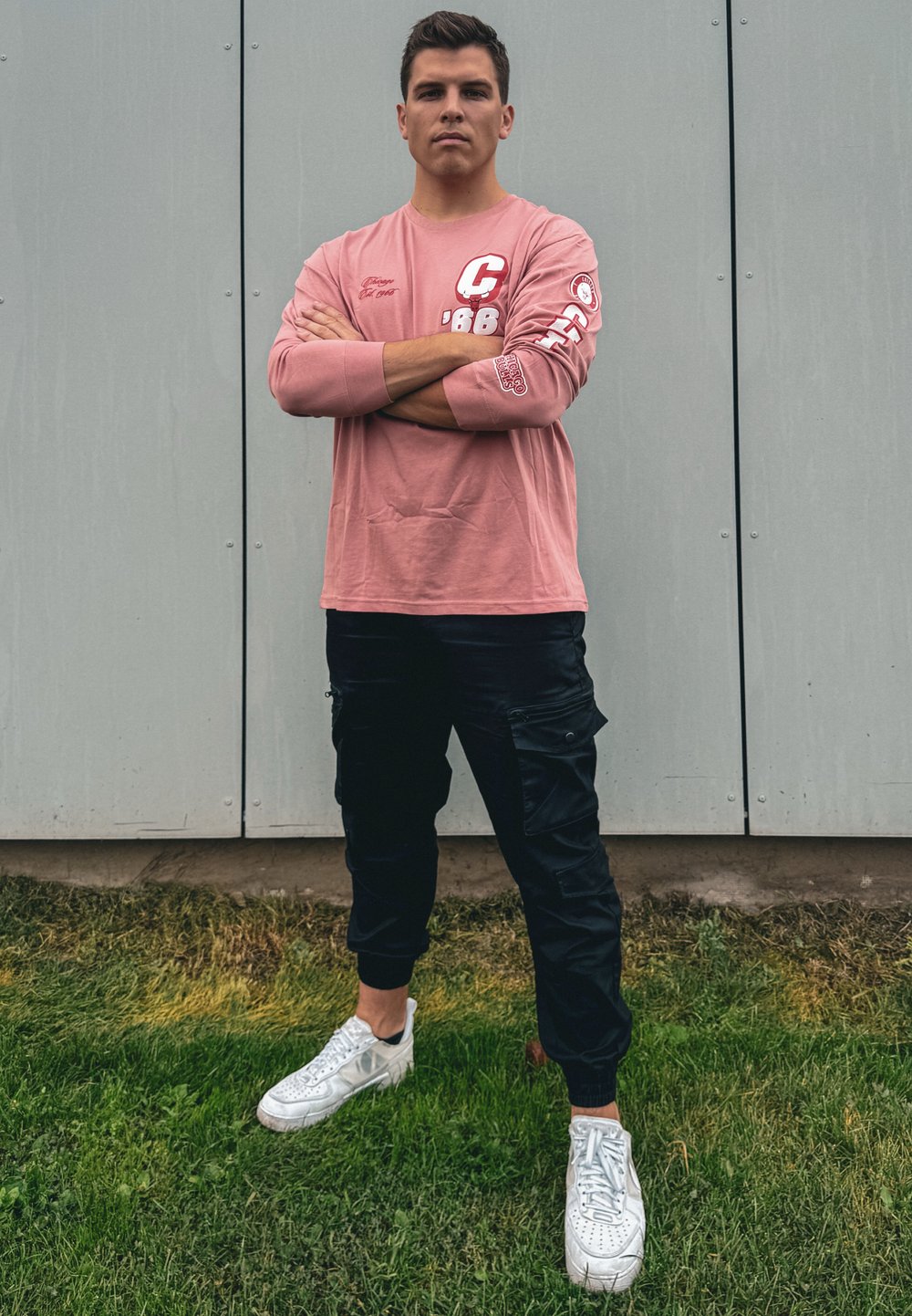 Νέος άντρας στέκεται σε γρασίδι με τα χέρια σταυρωμένα, φορώντας ροζ μακρυμάνικο πουκάμισο, μαύρο παντελόνι cargo και λευκά αθλητικά παπούτσια μπροστά σε γκρι πάνελ.