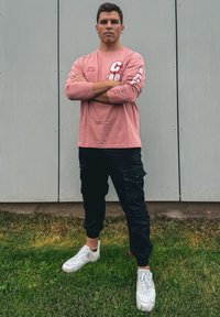 Νέος άντρας στέκεται σε γρασίδι με τα χέρια σταυρωμένα, φορώντας ροζ μακρυμάνικο πουκάμισο, μαύρο παντελόνι cargo και λευκά αθλητικά παπούτσια μπροστά σε γκρι πάνελ.