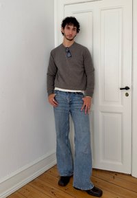 Grå strikket sweater over en hvid skjorte, vidde blå jeans og mørke sko. Krøllet hår med solbriller hvilende på sweateren.