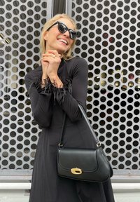 Črna obleka z dolgimi rokavi, ki vključuje črne perjanice na manšetah, v kombinaciji s črno torbico z zlatim zaklopom, postavljena na pikčasti ozadju.